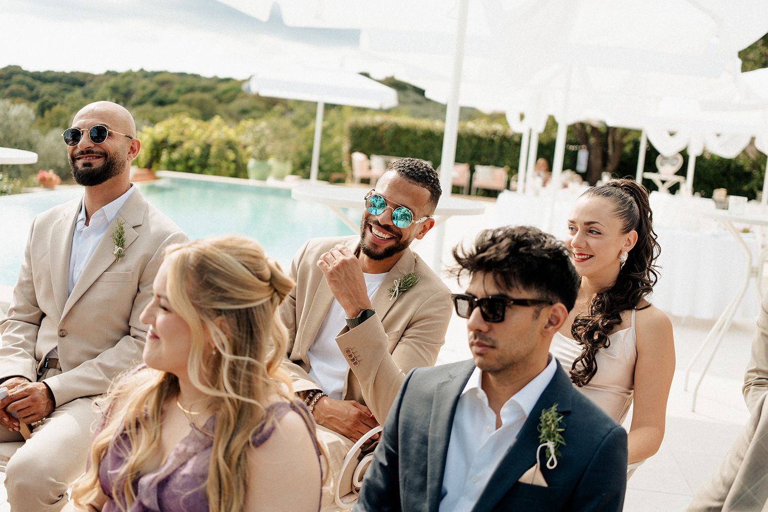 Guests in elegant attire smile and laugh at an outdoor wedding ceremony beside a pool on a sunny day.