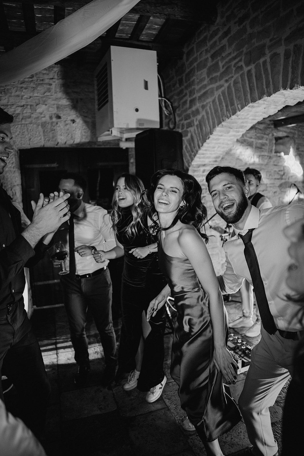 Black and white photo of joyful wedding guests dancing and celebrating at a rustic stone venue reception party.