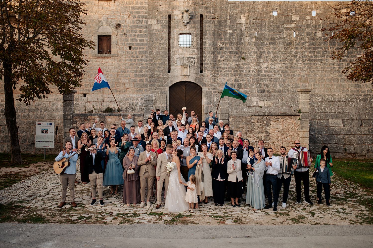Large wedding group photo in front of a historic stone castle wall with Croatian flags, surrounded by autumn trees.