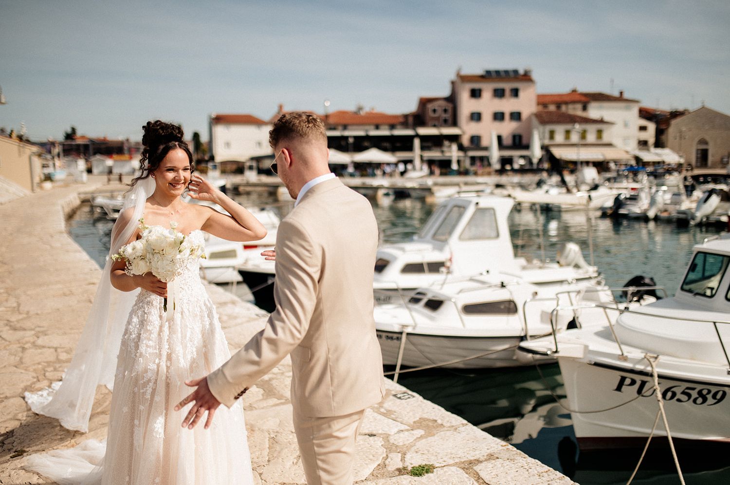 Bride and groom pose on a sunny harbor waterfront in Croatia, with white boats and historic buildings in the background.