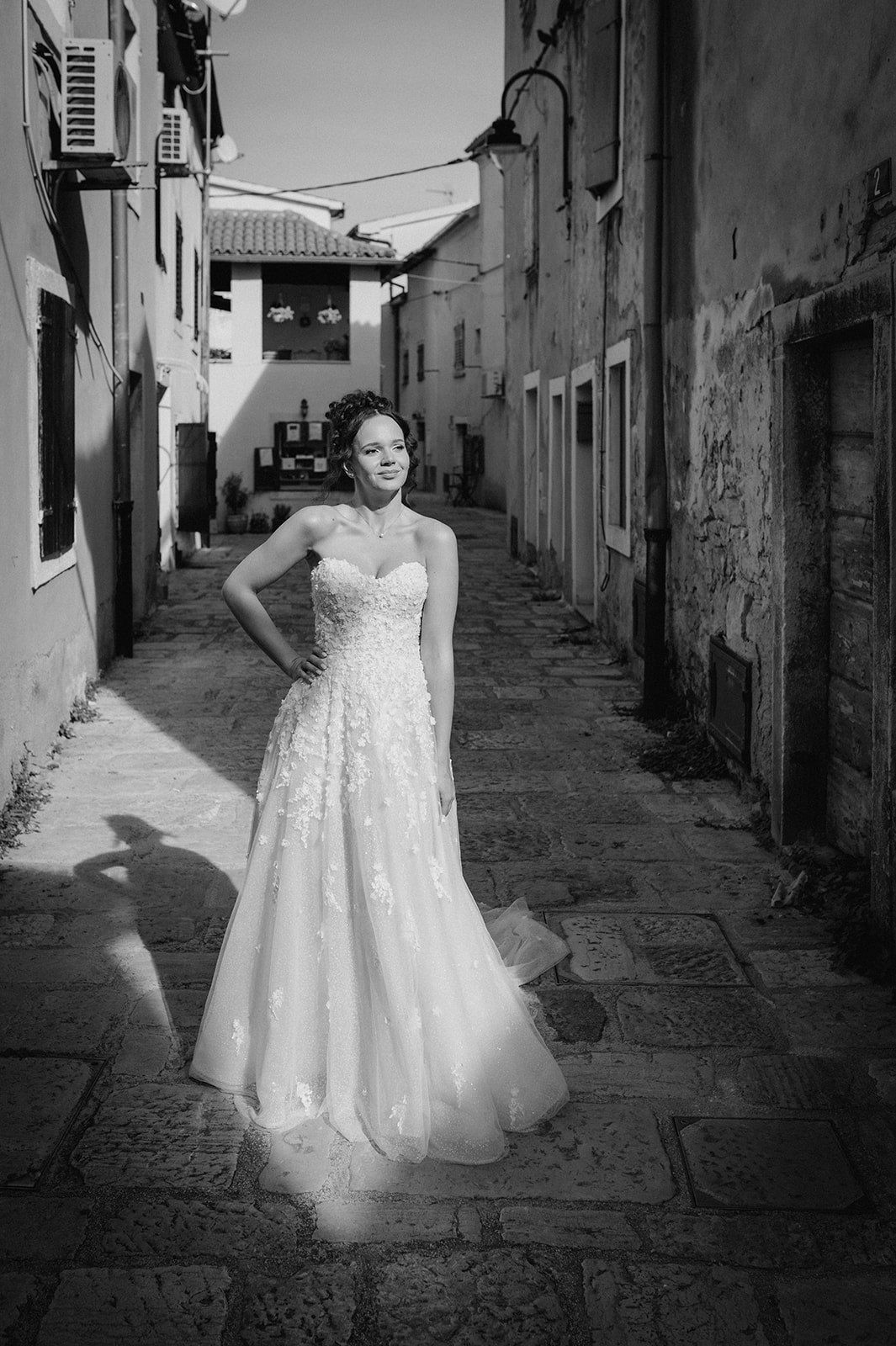Bride in strapless lace wedding gown poses in a sunlit cobblestone alley between old stone buildings.
