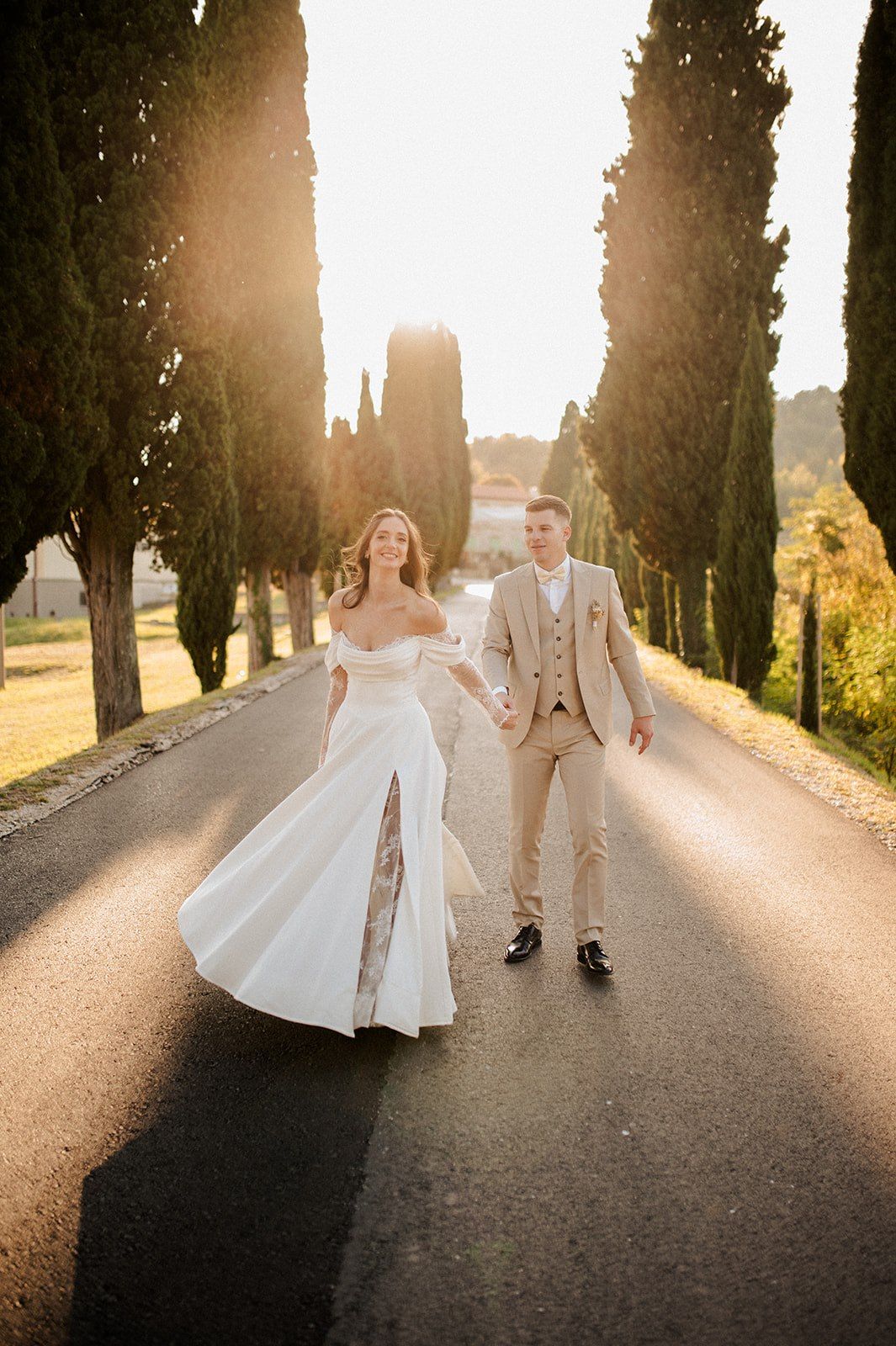 Bride and groom walk hand-in-hand down a sun-drenched cypress-lined road in Tuscany.