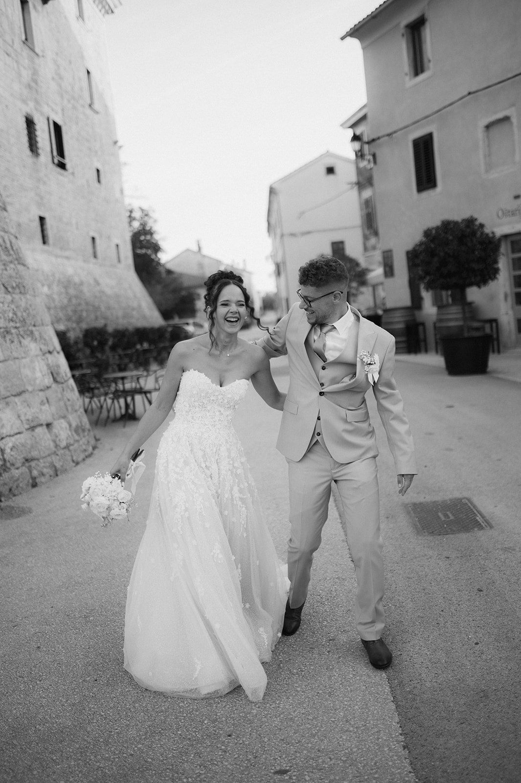 Joyful bride and groom walking together on a cobblestone street in a black and white wedding photo.