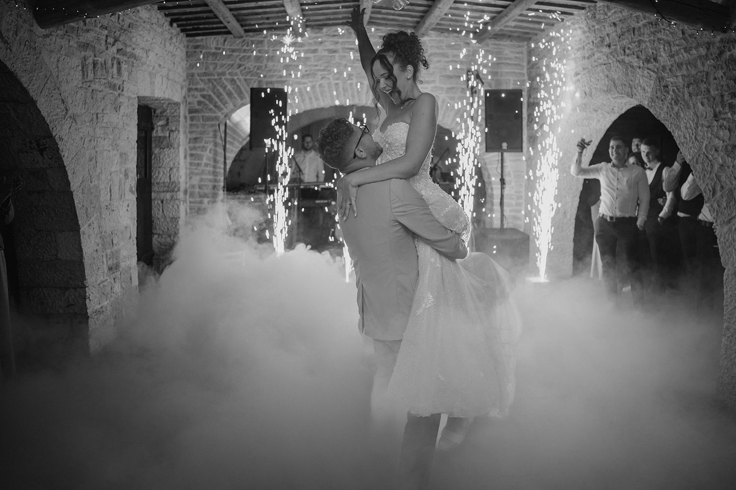 Black and white wedding first dance photo with dramatic fog effect in rustic stone venue with fairy lights.