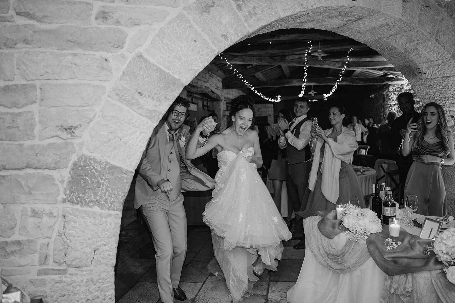 Bride and groom dancing through a stone archway surrounded by cheering guests at a wedding reception.