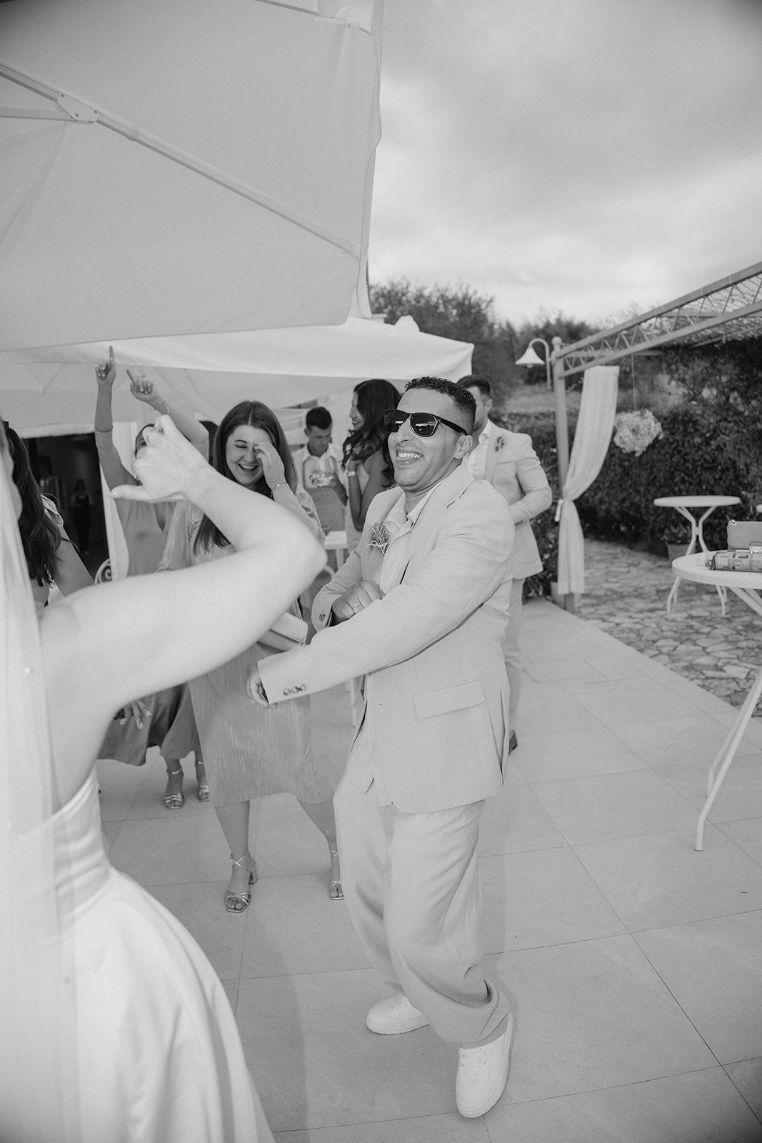 Black and white photo of joyful groom in sunglasses dancing with guests at outdoor wedding.