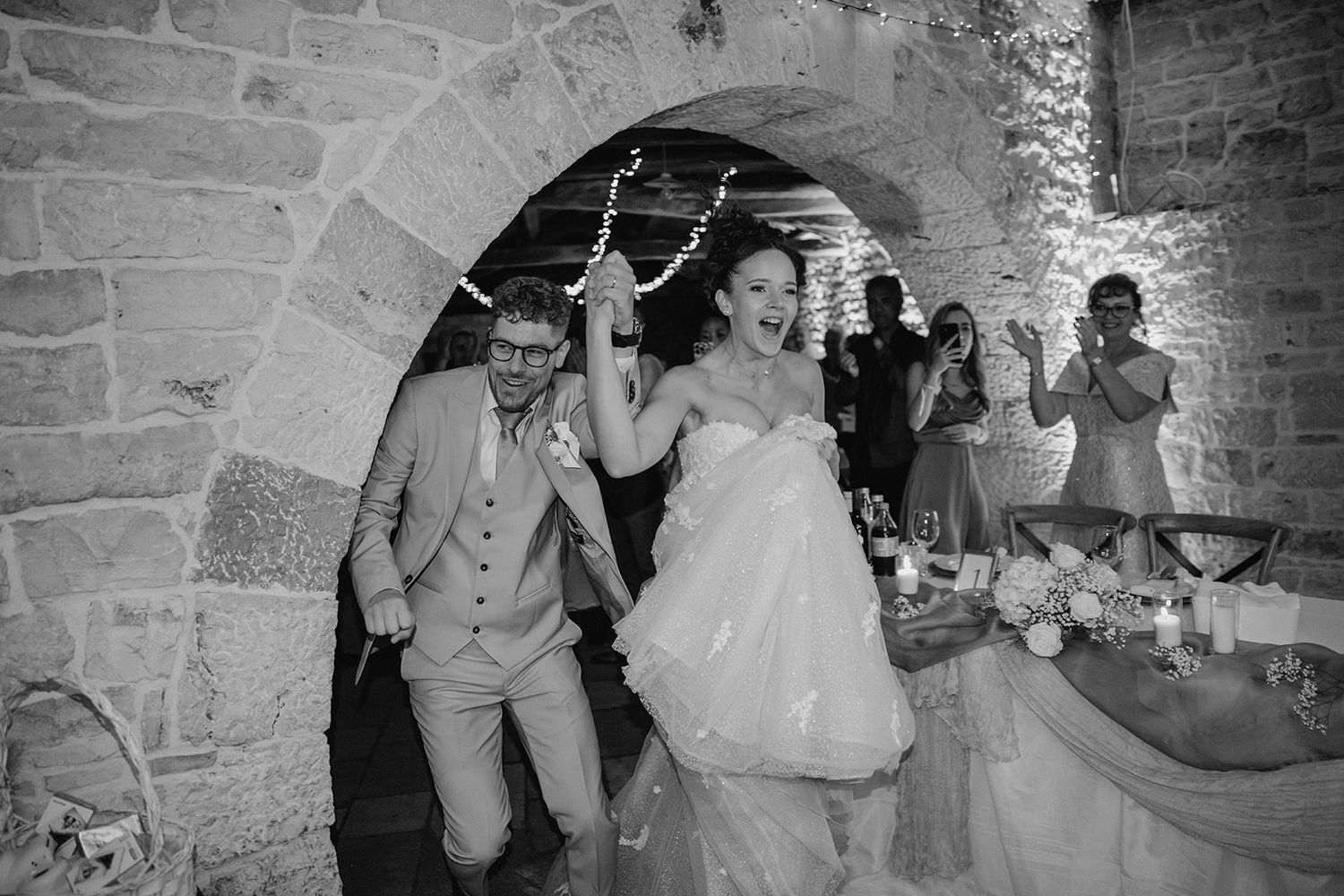 Black and white photo of a joyful bride and groom making a grand entrance through a stone archway at their wedding.