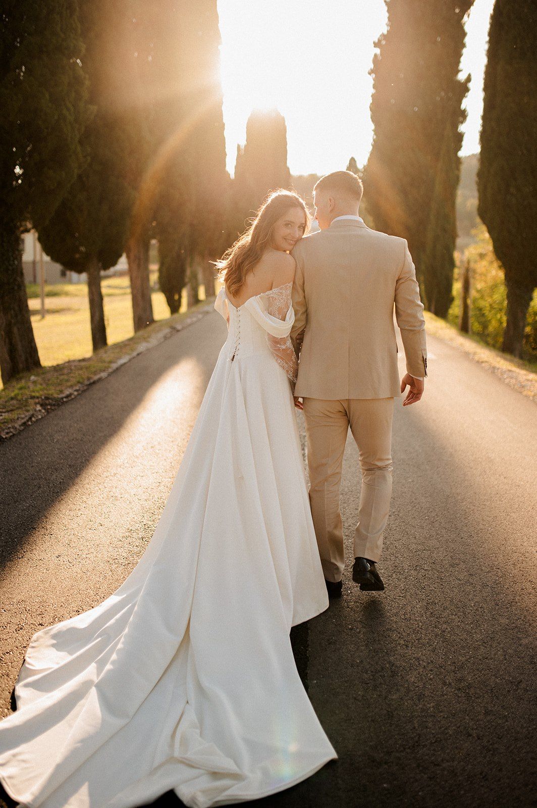 Bride in white gown and groom in beige suit walk down tree-lined road at golden sunset.