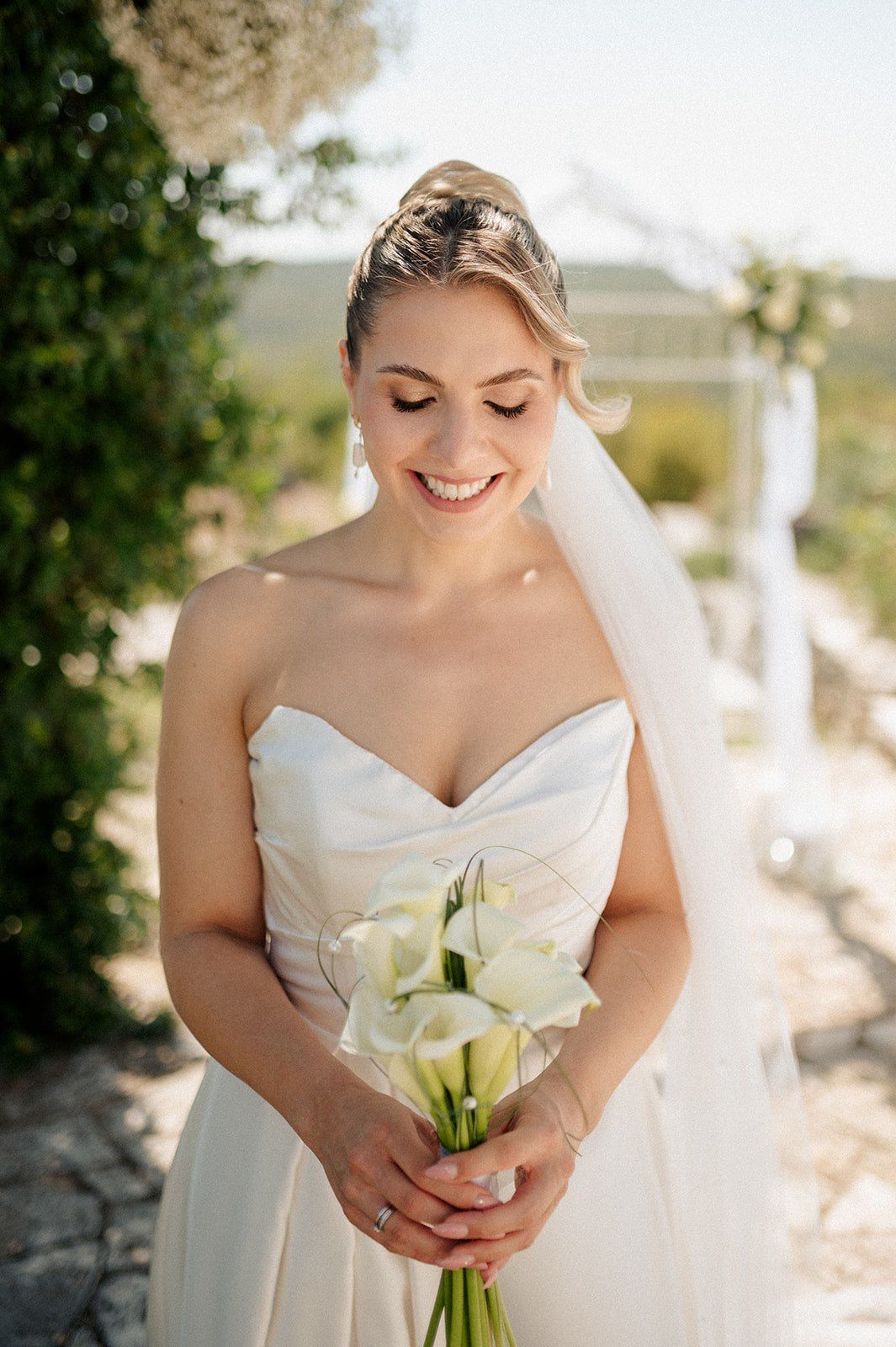 Smiling bride in strapless white gown holds calla lily bouquet outdoors on sunny wedding day.