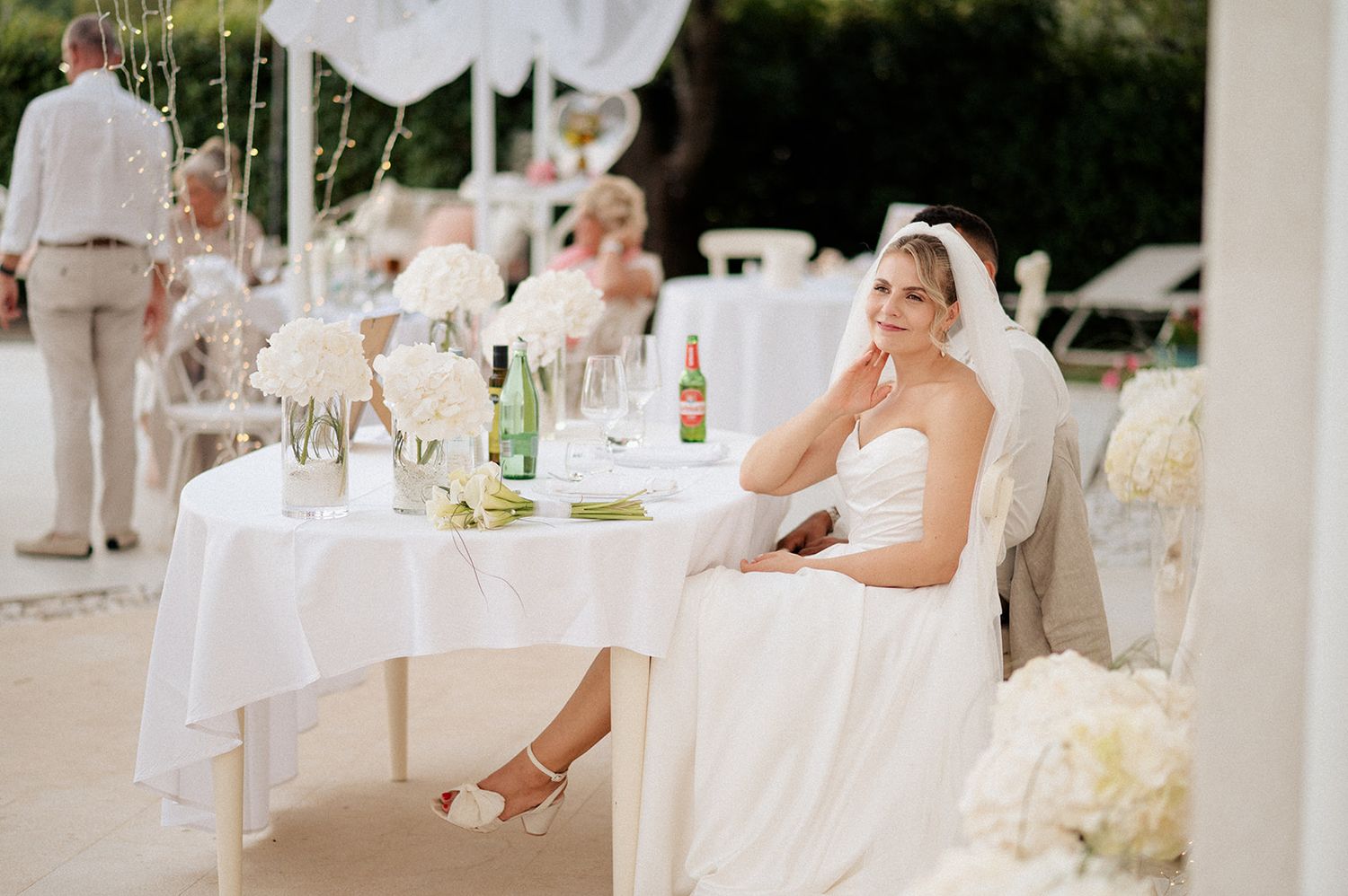 Elegant bride in white strapless gown seated at decorated wedding reception table with floral arrangements.