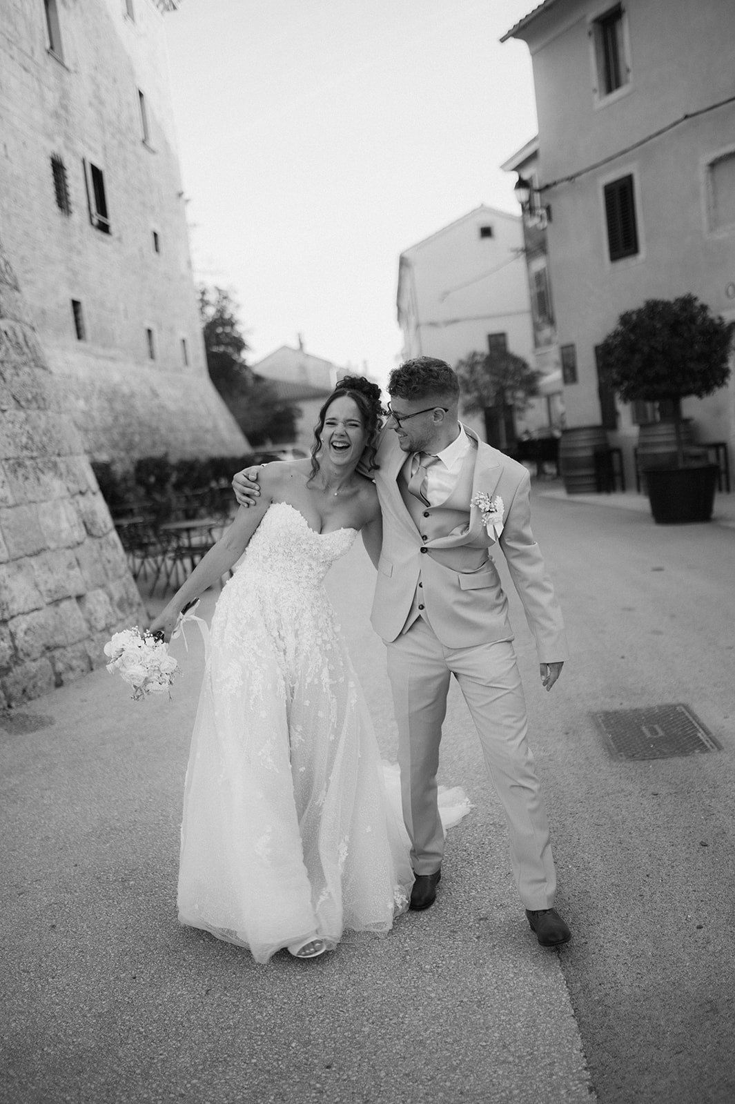 Bride and groom laughing together on a cobblestone street in a black and white wedding photo.