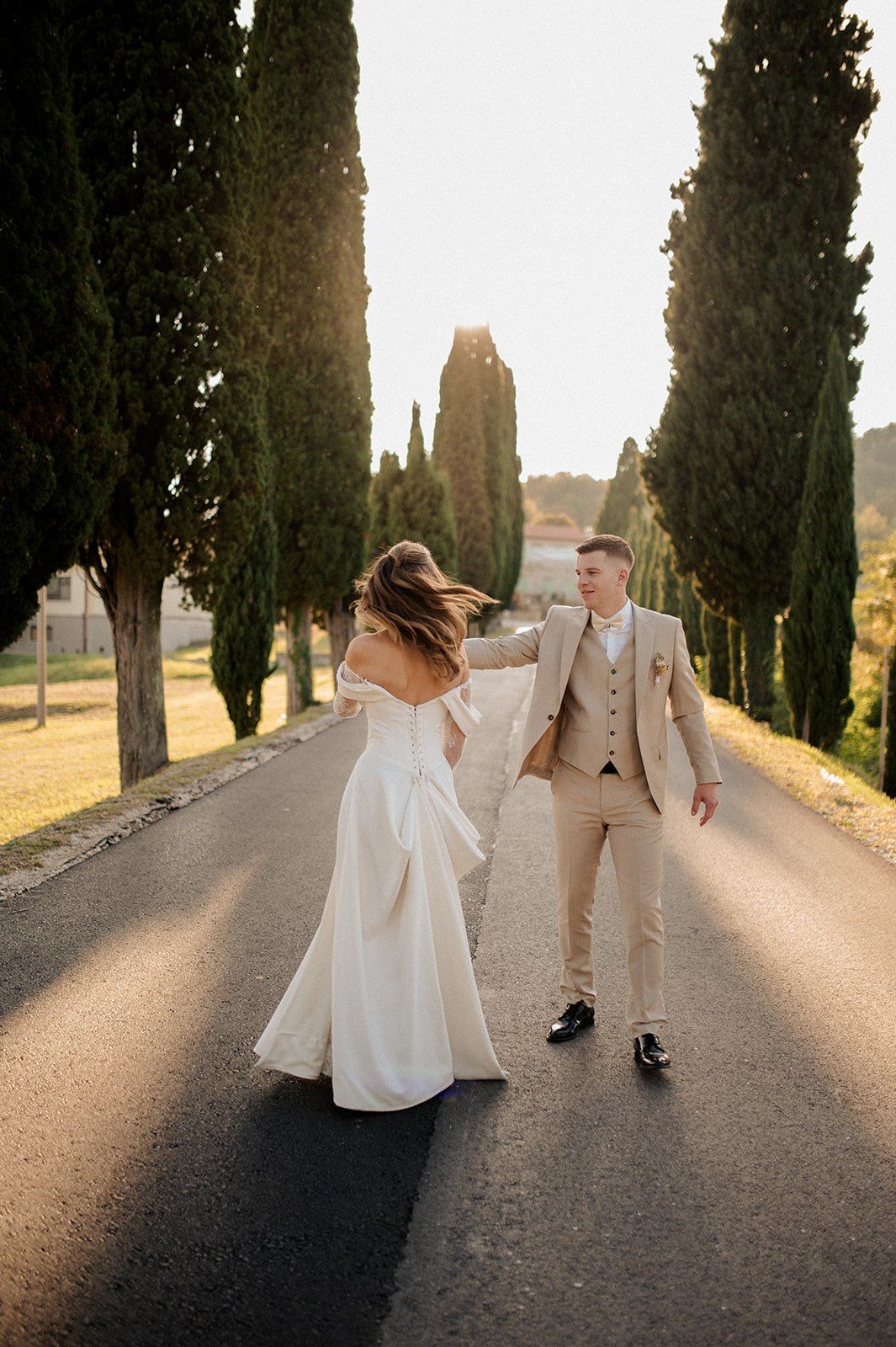 Bride and groom dancing on a tree-lined road at golden hour, bride in white off-shoulder gown, groom in beige suit.