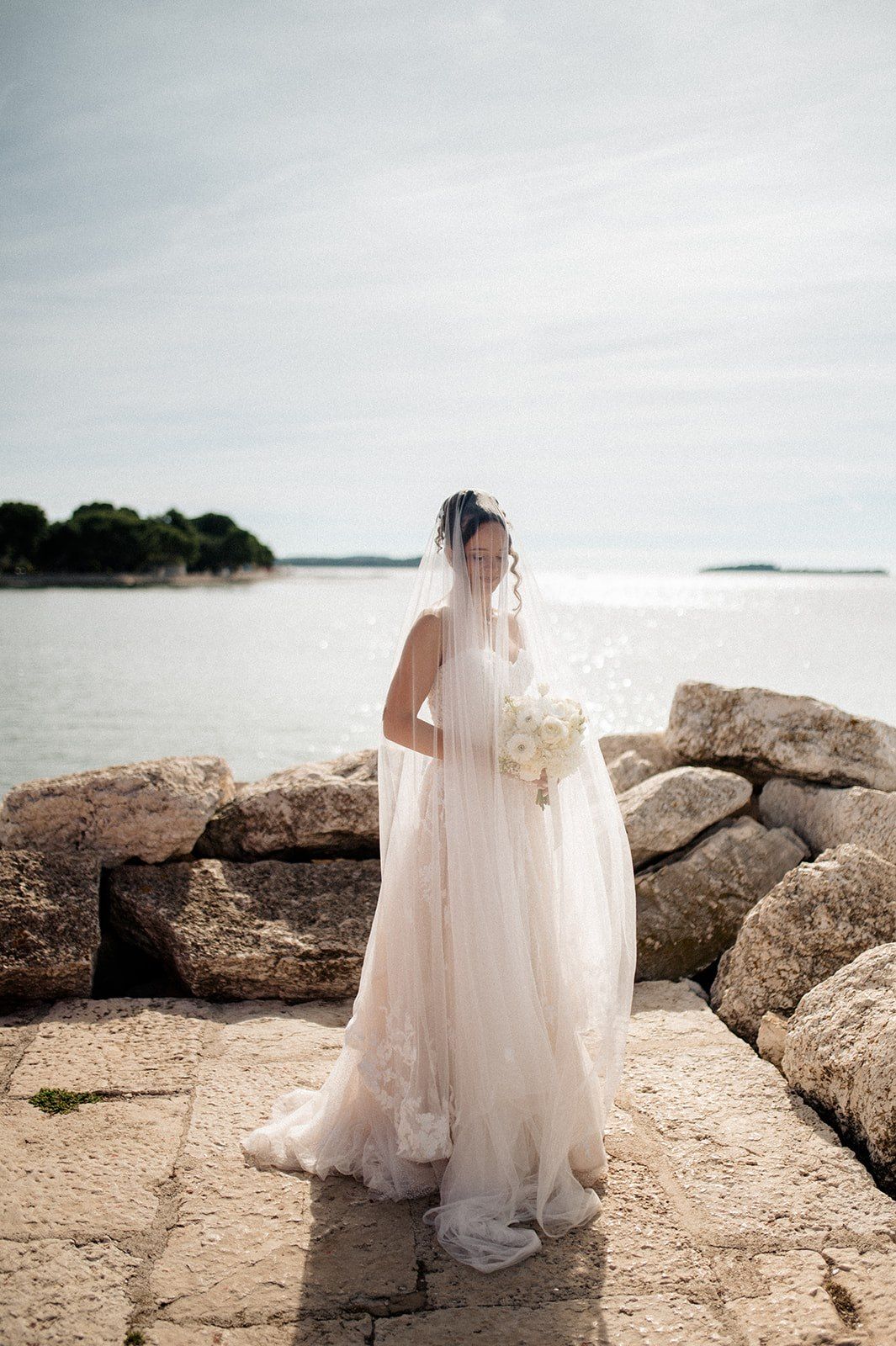 Bride in white lace gown and veil holds bouquet on rocky shore with serene ocean backdrop.