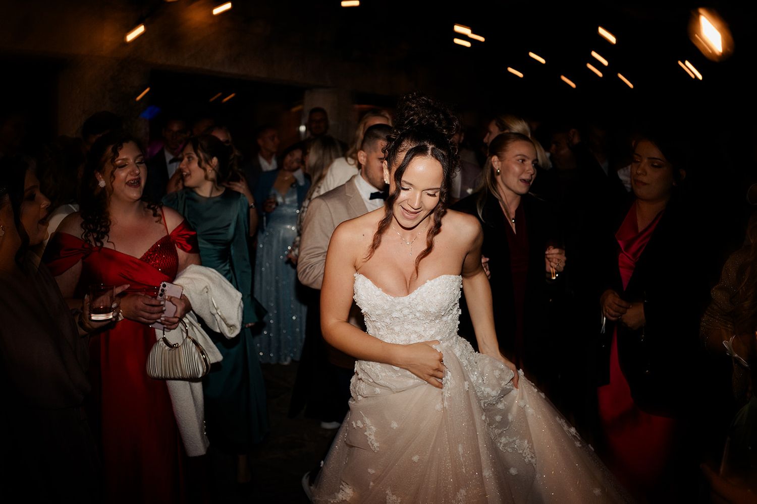 Bride in a strapless lace wedding gown dancing joyfully with guests at a lively wedding reception.