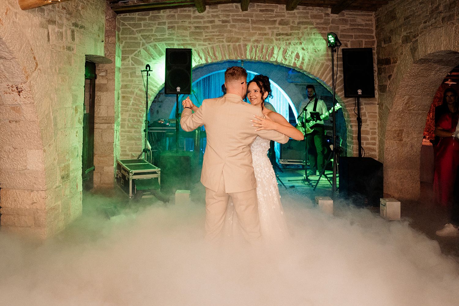 Bride and groom share first dance on fog-covered floor at rustic stone venue with live band performing in background.