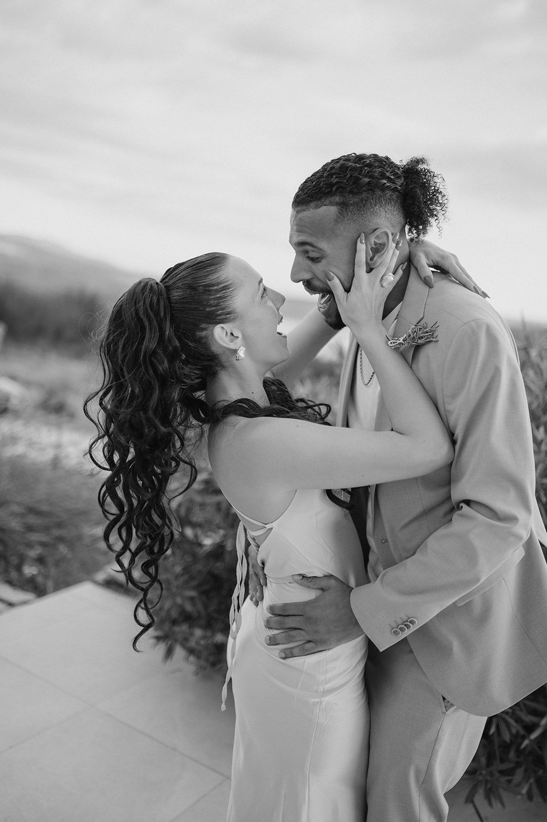 Black and white photo of joyful newlywed couple embracing and laughing together after outdoor wedding.