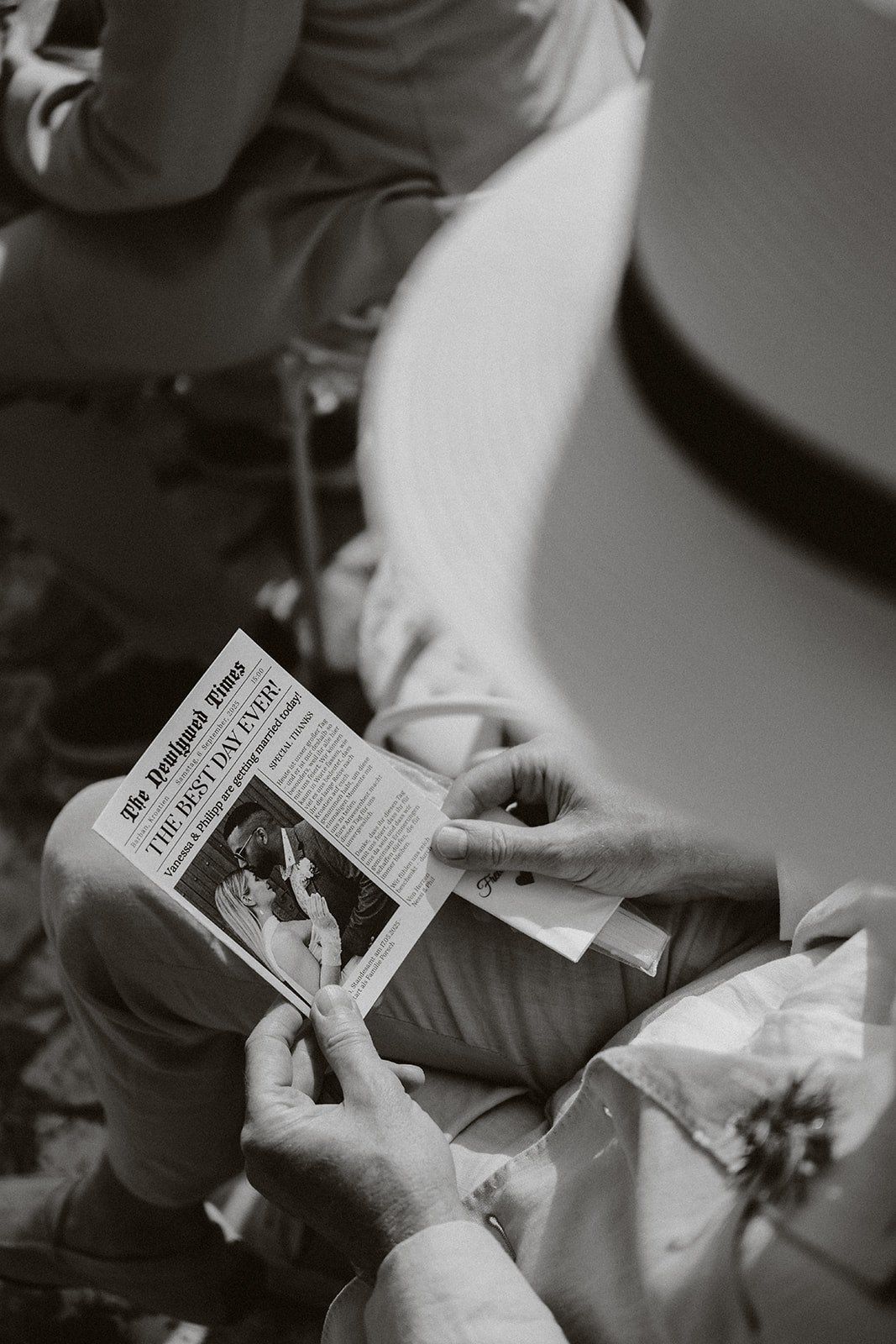 Black and white photo of hands holding and reading a small printed newspaper or pamphlet while sitting.