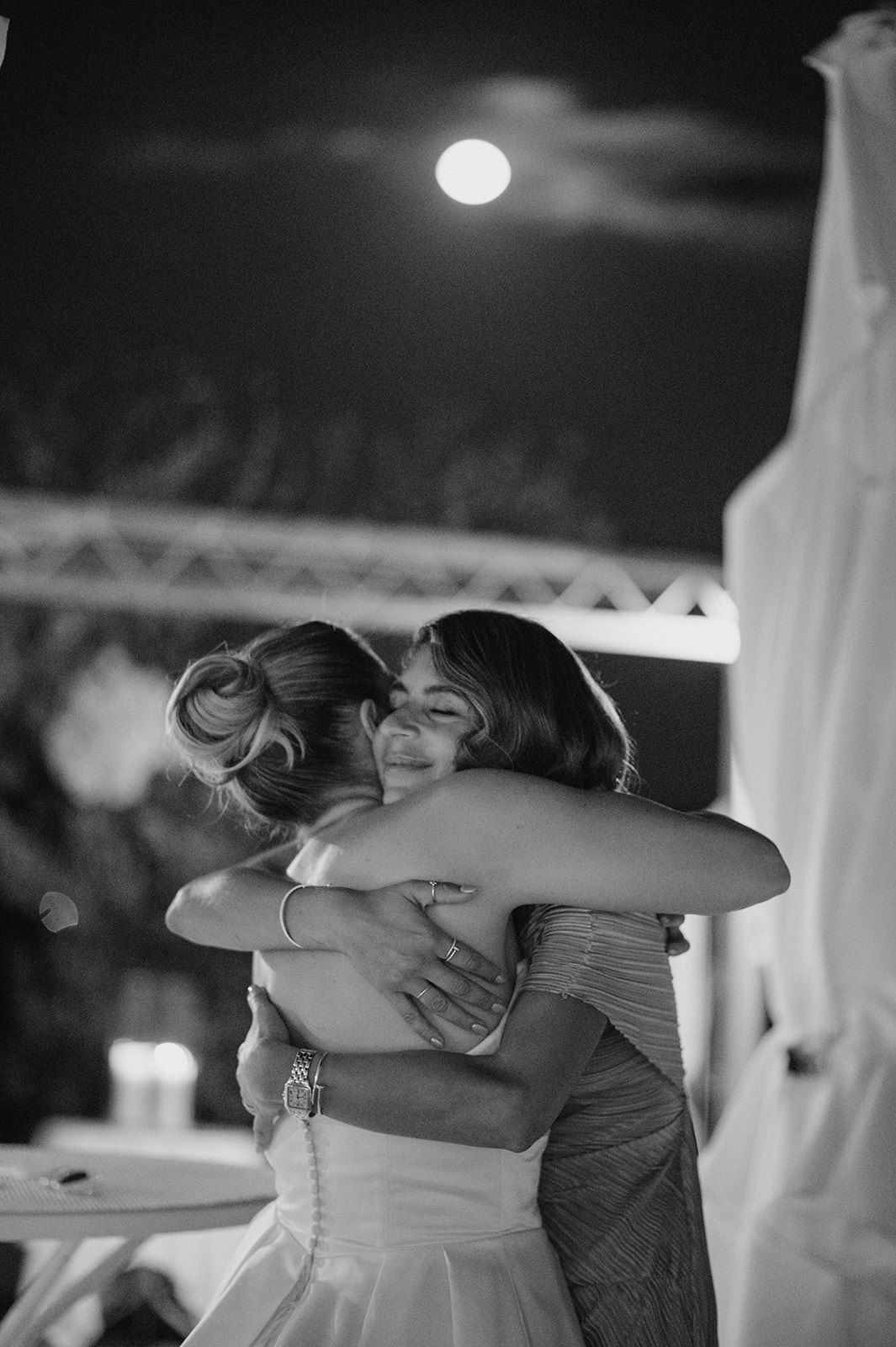 Bride and guest share an emotional hug under a full moon at an outdoor wedding reception in black and white.
