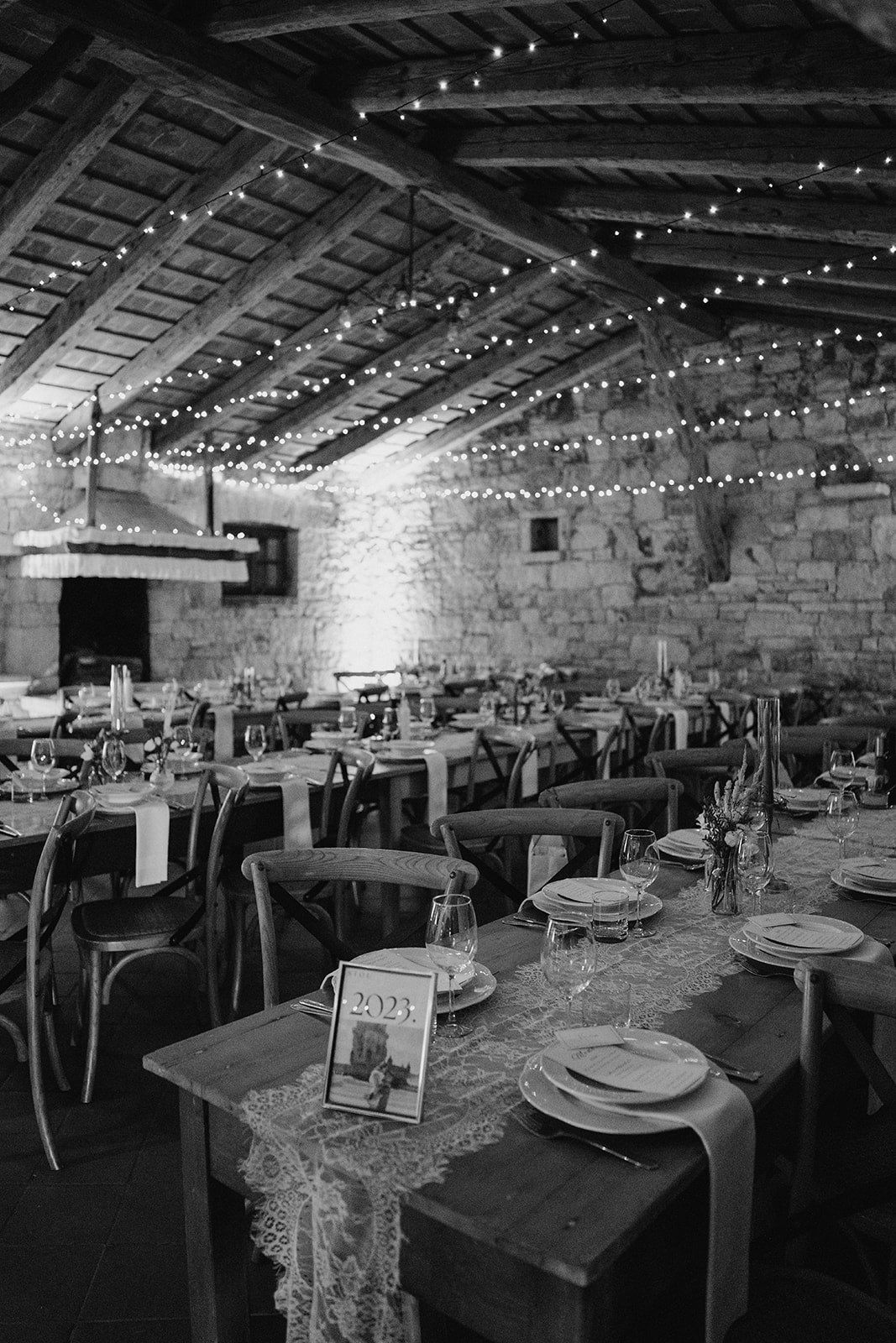 Black and white rustic barn wedding reception with string lights, wooden tables, cross-back chairs, and elegant place settings.