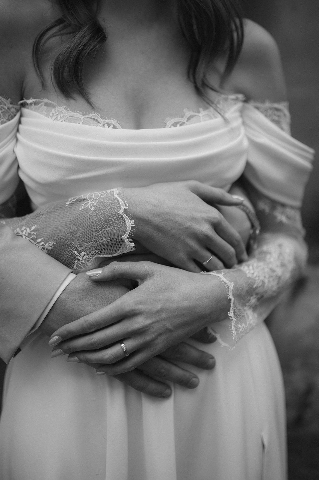 Black and white photo of a couple embracing, showing wedding rings on their hands, bride in elegant lace off-shoulder gown.