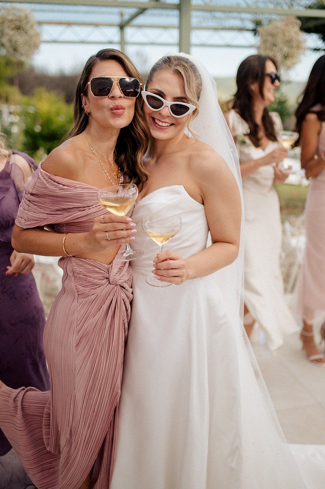 Bride in white strapless gown and veil poses with bridesmaid in pink dress, both wearing sunglasses and holding champagne.