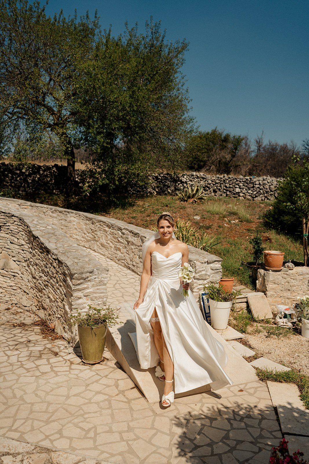 Bride in white strapless gown holding bouquet walks stone path in sunny Mediterranean garden setting.