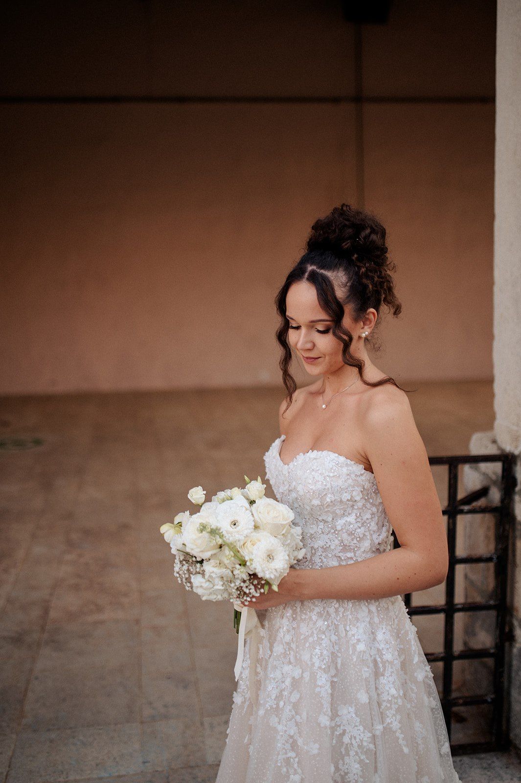 Bride in strapless lace wedding gown holding white rose bouquet, looking down gracefully at outdoor venue.
