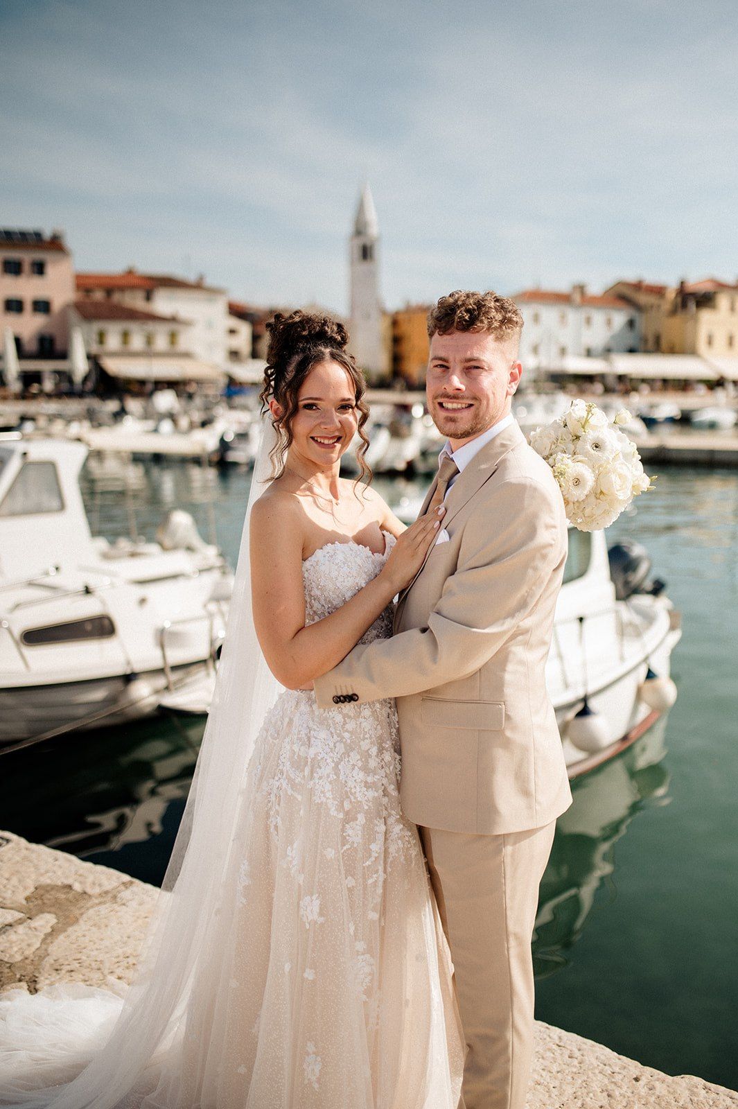 Bride and groom pose at a Mediterranean harbor with boats and a church tower, bride in lace gown holding white bouquet.