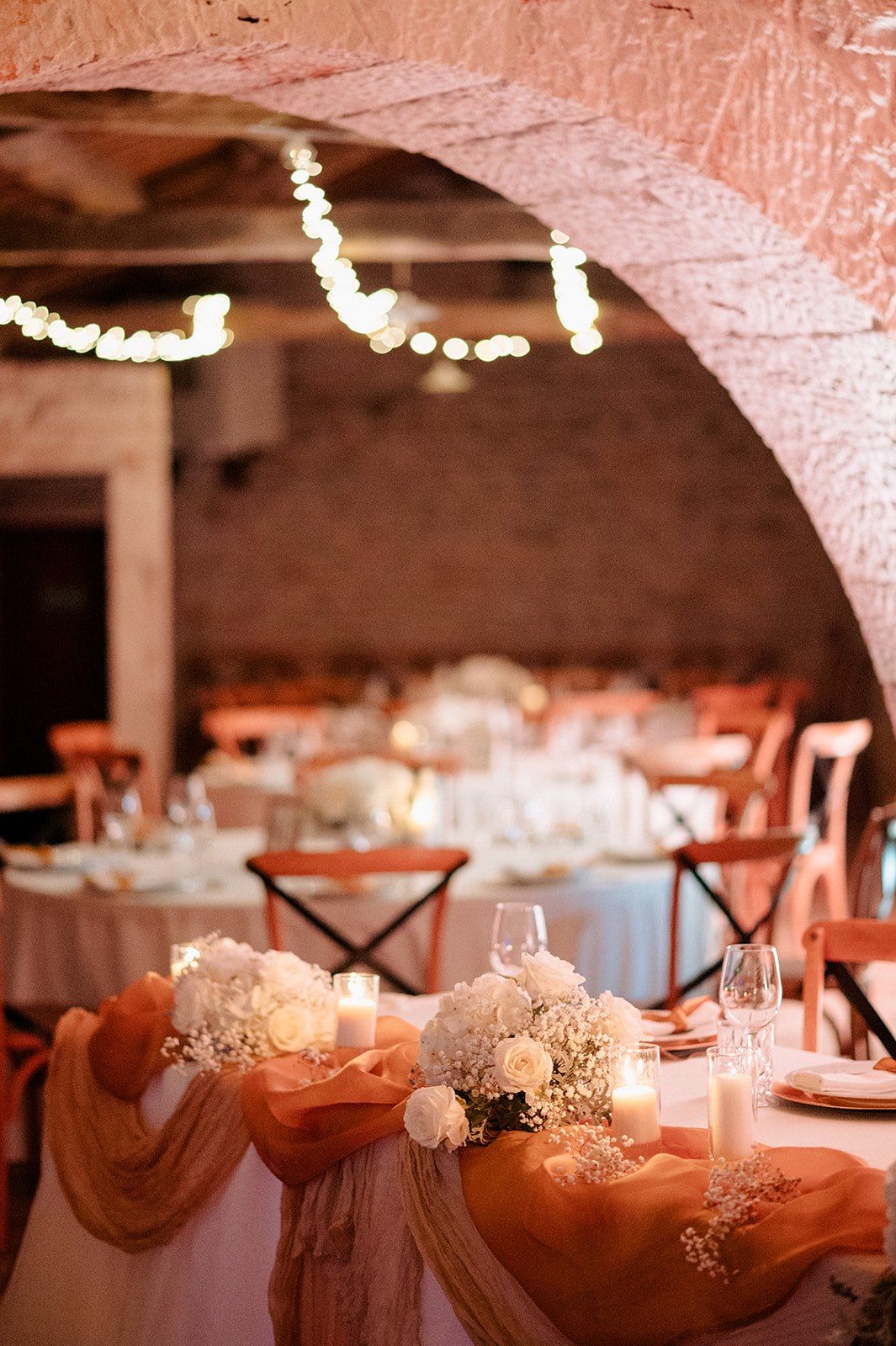 Elegant wedding reception with white floral centerpieces, candles, peach drapery, and string lights under a stone arch.