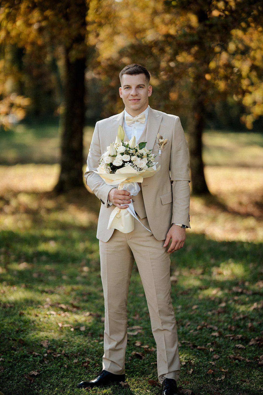 Groom in beige suit holding white flower bouquet standing in autumn park with golden trees.