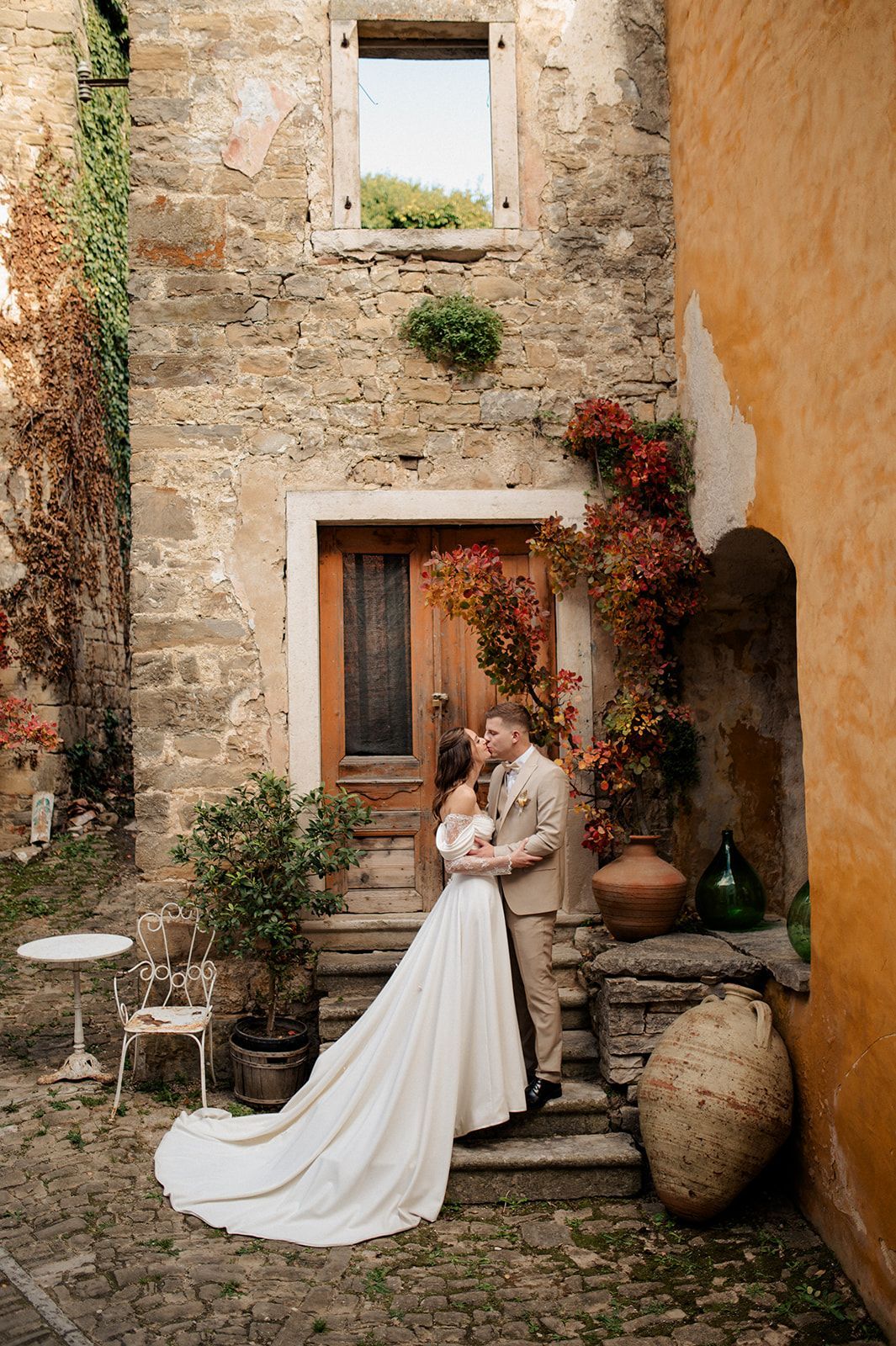 Bride and groom embrace in a romantic Italian courtyard with stone walls, red foliage, and rustic wooden door.