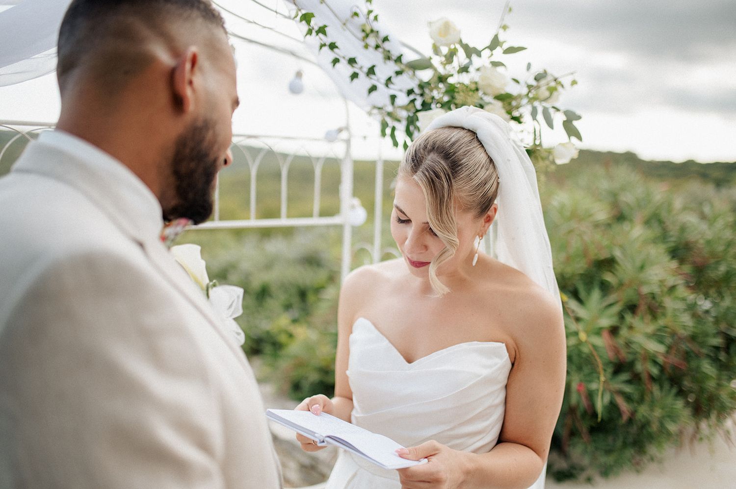 Bride reads wedding vows to groom during outdoor ceremony with floral arch backdrop.