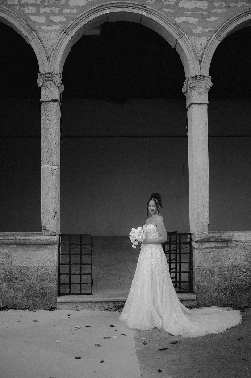Bride in a white gown holding a bouquet stands under stone arches in a black and white wedding photo.
