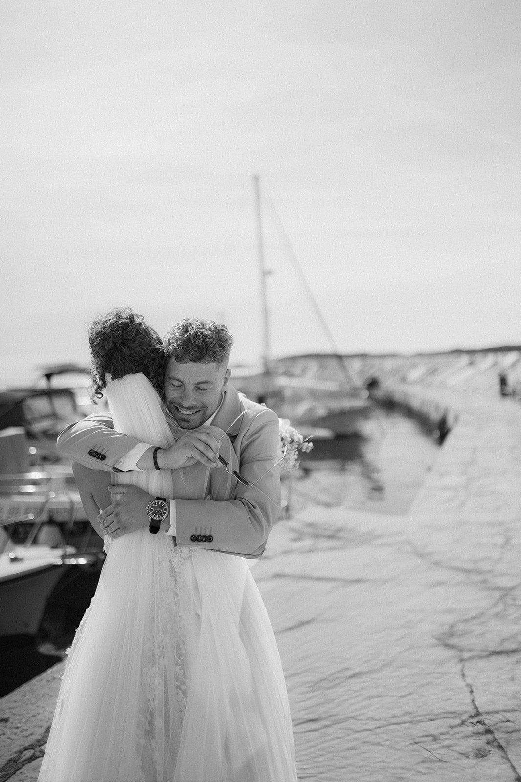 Black and white photo of a smiling groom hugging his bride near a marina with boats in the background.