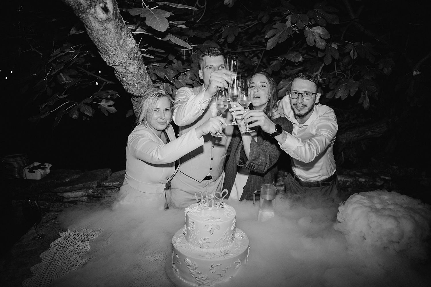 Black and white photo of wedding party toasting with champagne glasses around a decorated cake outdoors at night.