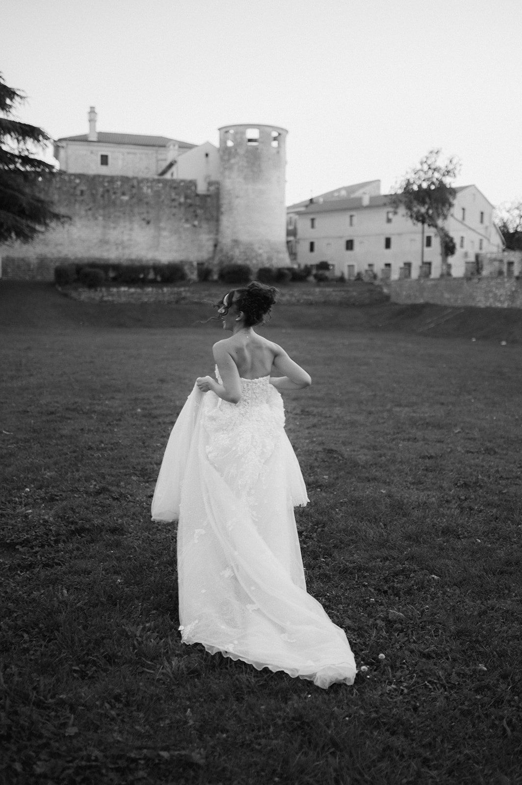 Black and white photo of a bride in a flowing wedding gown walking toward a historic stone castle on a green lawn.