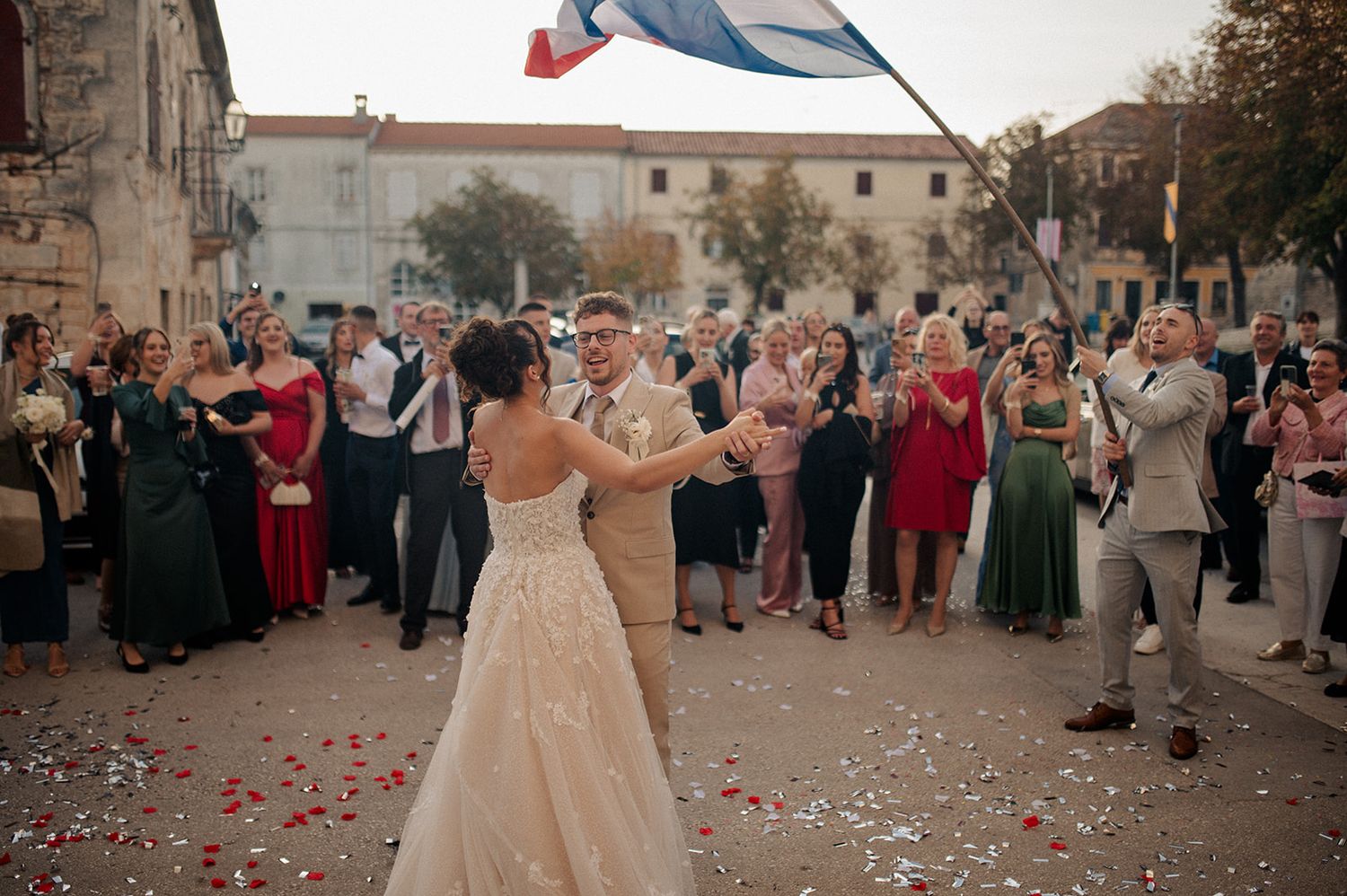 Bride and groom dancing outdoors at a wedding celebration with guests cheering and confetti in a European town square.
