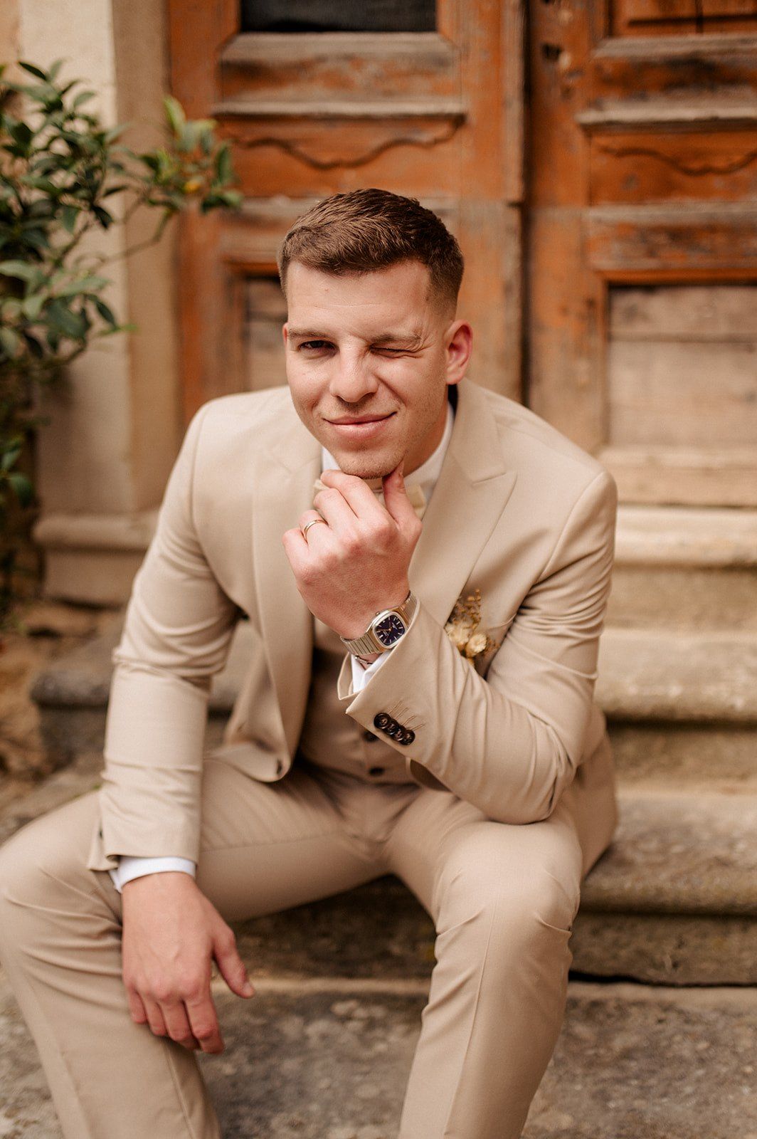 A stylish groom in a beige suit sits on stone steps, smiling thoughtfully while touching his chin near a wooden door.