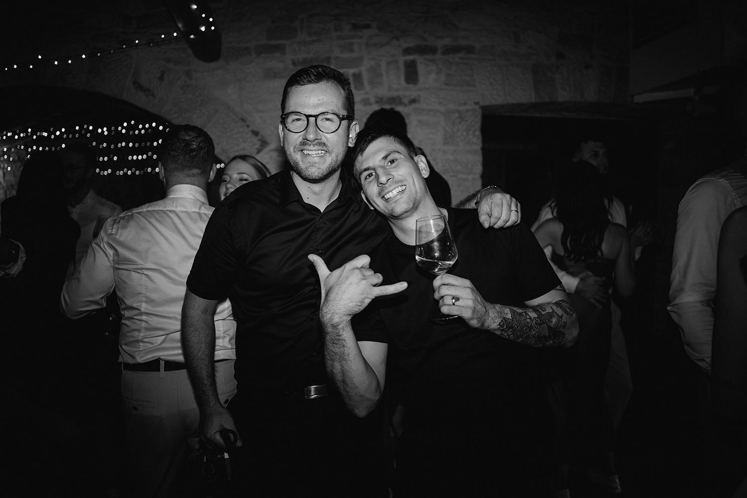 Two men in black shirts smiling and posing together at a lively wedding reception party.