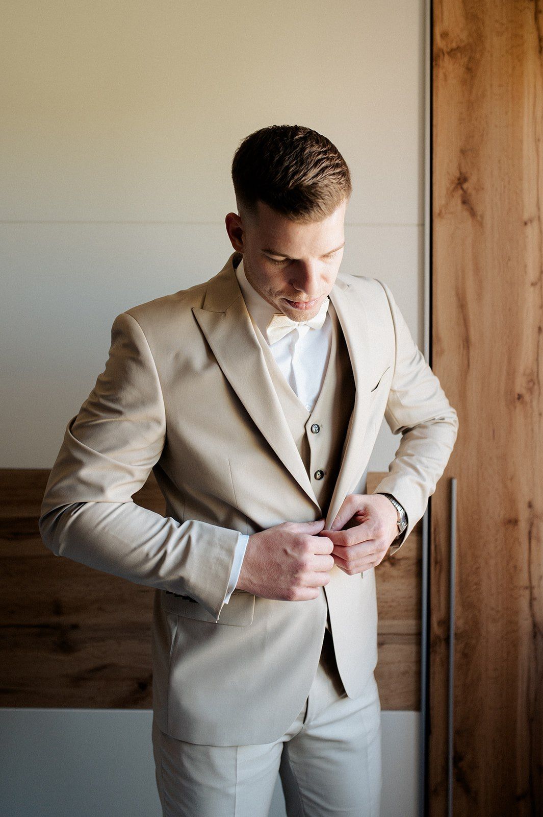 A groom in a beige three-piece suit adjusts his jacket button while preparing for his wedding day.