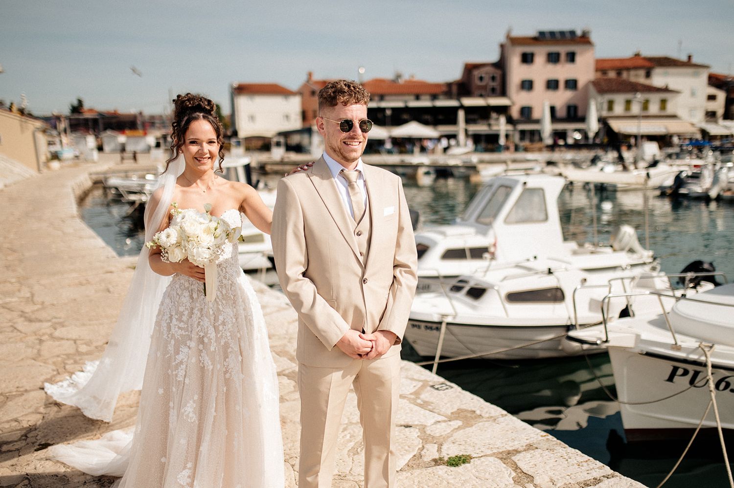 Bride in lace gown and groom in beige suit pose by a sunny Mediterranean harbor with boats and stone buildings.