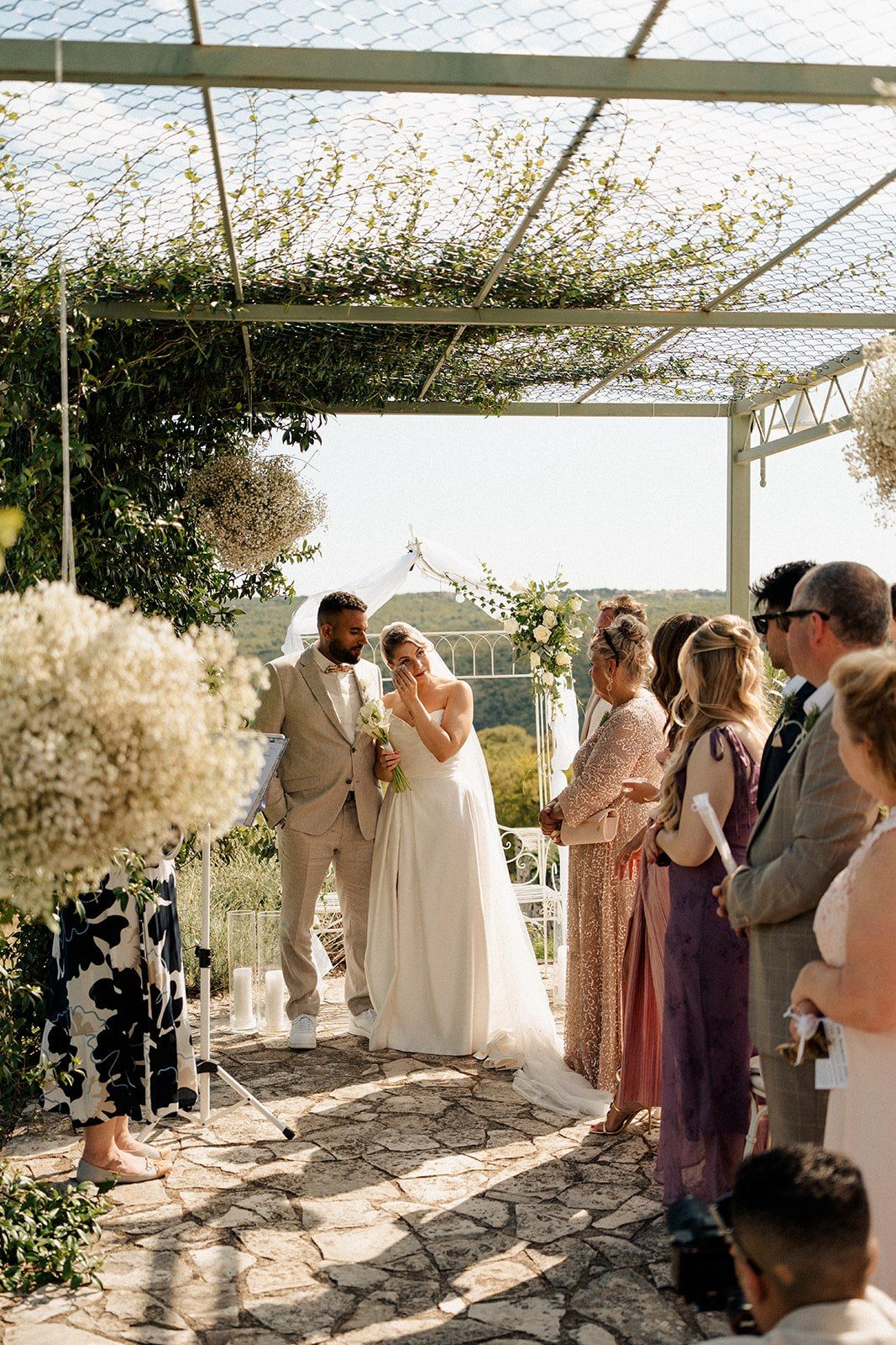 Couple shares a romantic moment during an outdoor wedding ceremony under a floral pergola with guests watching.