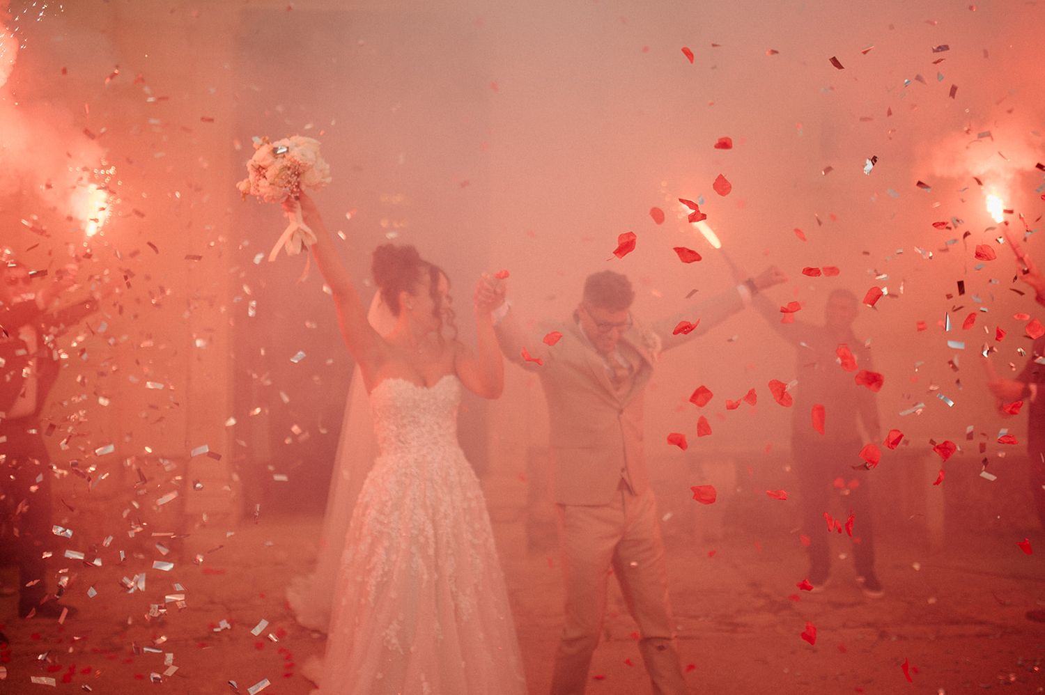 Bride and groom celebrate with red confetti and flares at dramatic wedding reception entrance.