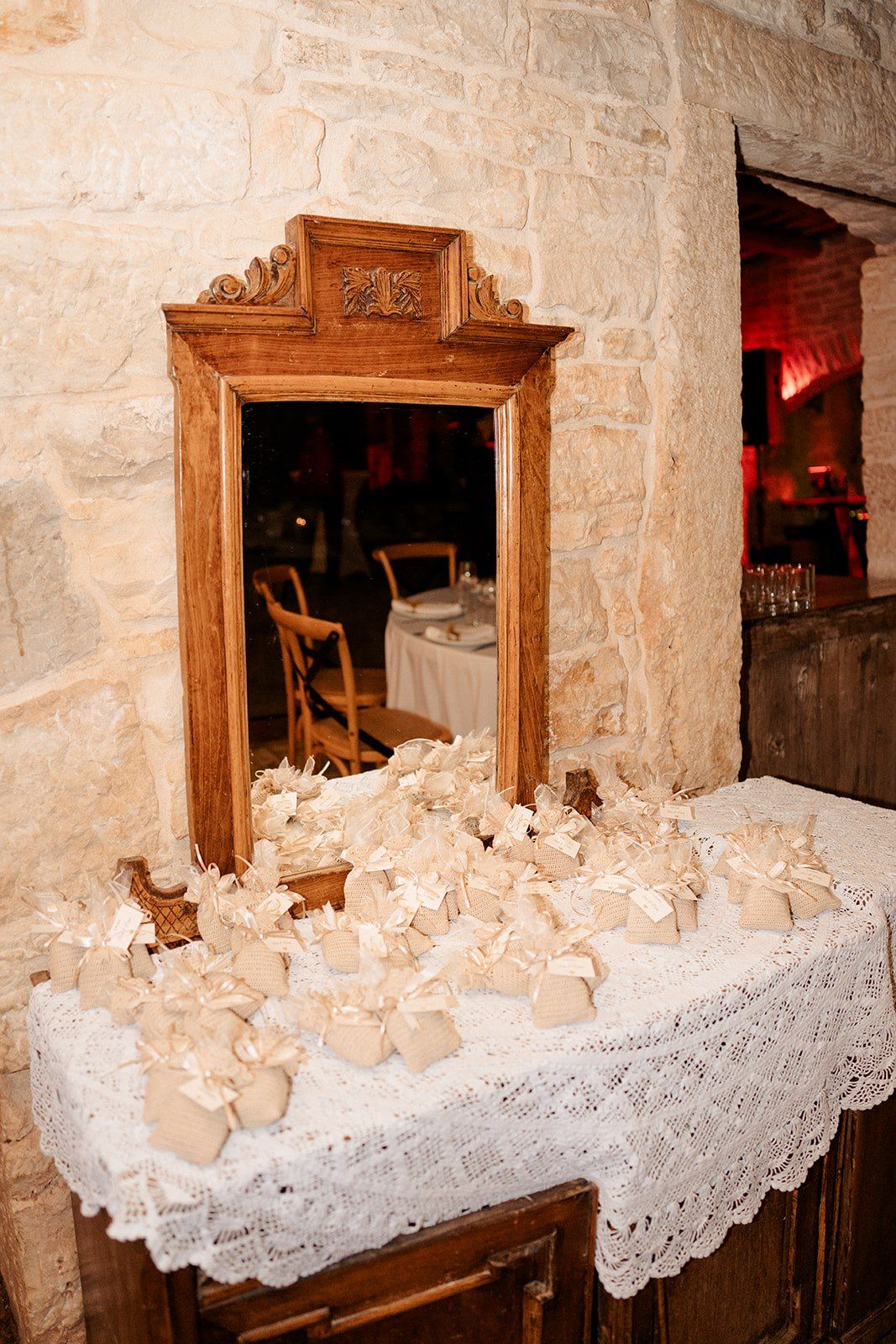 Elegant wedding favor table with lace cloth, ivory gift boxes, and ornate wooden mirror against stone wall.