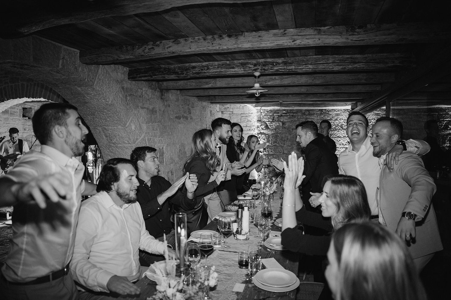 Guests laughing and celebrating at a rustic stone wedding reception dinner in black and white photography.