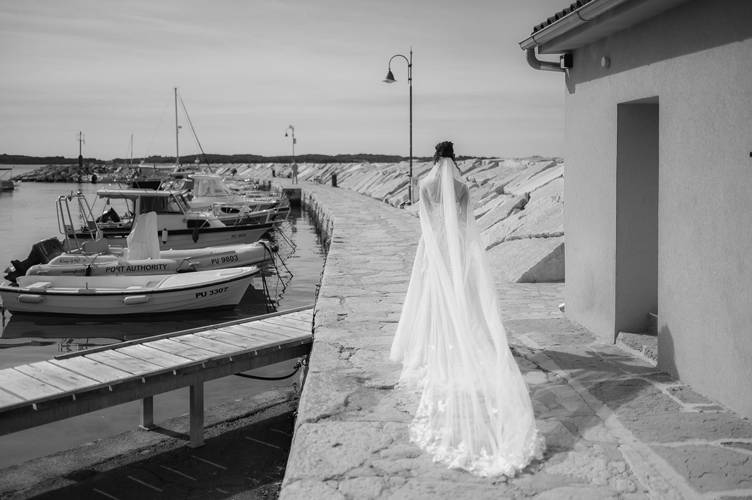 Bride in white gown and veil stands on stone pier overlooking boats at harbor in black and white photo.