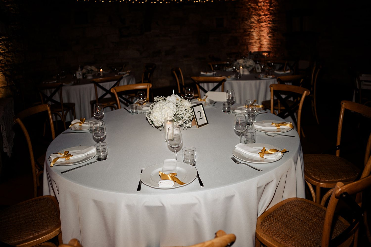 Elegant wedding reception table with white linen, floral centerpiece, gold accents, and glassware in a warmly lit venue.