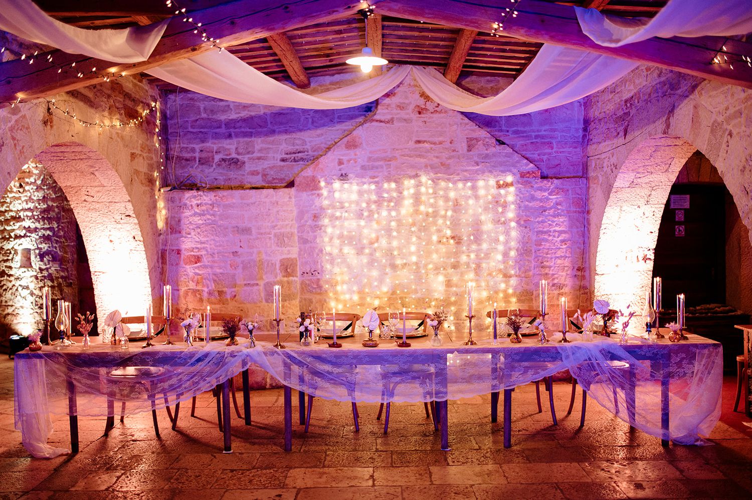 Elegant wedding reception in rustic stone venue with purple uplighting, fairy light backdrop, and draped fabric ceiling.