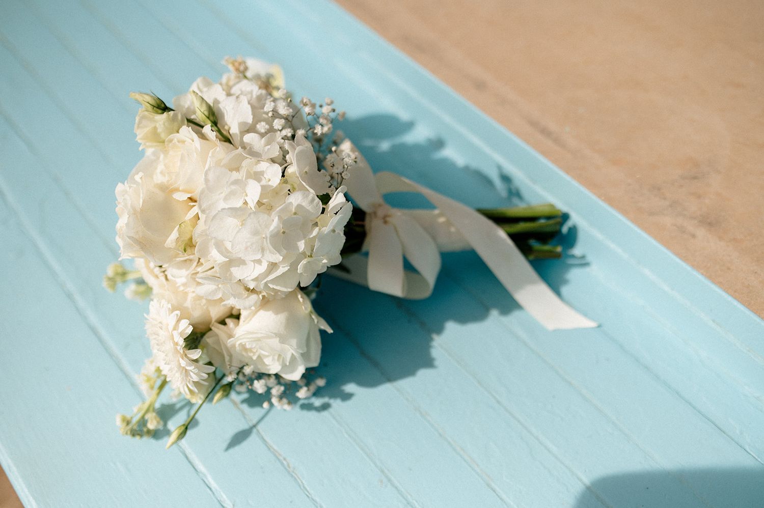 White peony and baby's breath bridal bouquet with ribbon on a light blue wooden surface.
