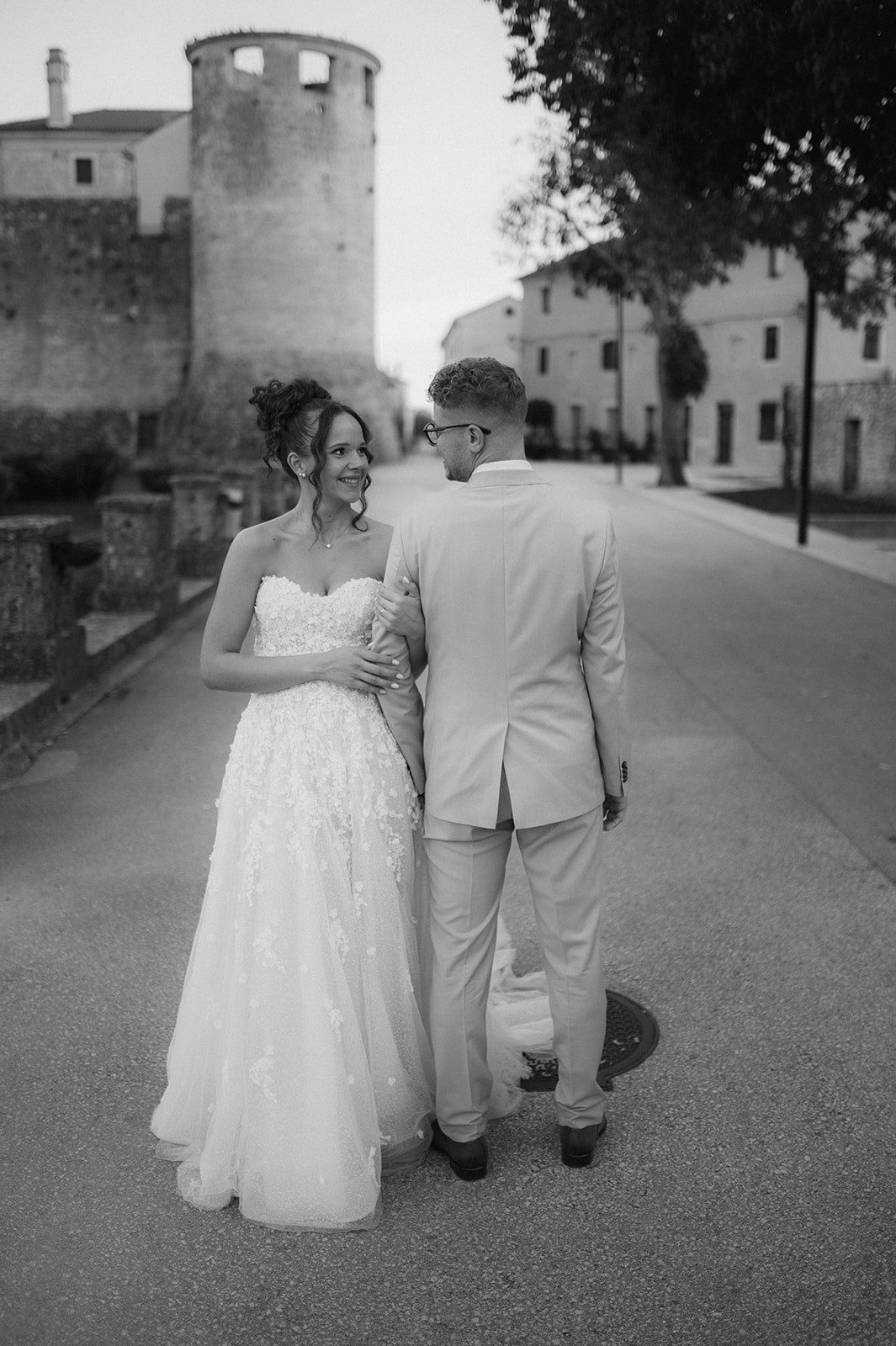 Black and white photo of a bride and groom walking together on a street, with historic buildings in the background.